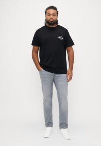 Μαύρο βαμβακερό t-shirt με κοντά μανίκια και λογότυπο μπροστά, συνδυασμένο με ανοιχτό μπλε τζιν παντελόνι και λευκά αθλητικά παπούτσια. Μινιμαλιστικός σχεδιασμός.