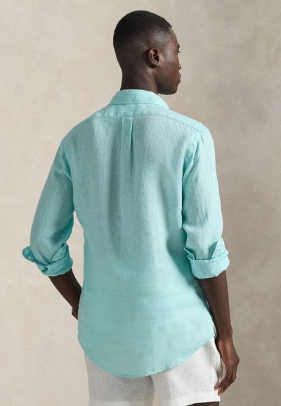 Ανοιχτό μπλε πουκάμισο λινό με μανίκια γυρισμένα, λεπτομέρεια στην πλάτη και στρογγυλεμένη βάση. Φορεμένο πάνω από λευκά σορτς. Λεία υφή και άνετη εφαρμογή.