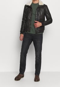 Brun læderjakke med lammeskindskrage, grøn t-shirt, mørke jeans og brune ankelstøvler. Slim fit-design med forlommer og lynlås.