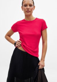 Ryškiai rožinė banguota trumpomis rankovėmis marškinėliai derinami su juoda klostuota sijonu, demonstruojančiu lygią medžiagą ir pritaikytą siluetą.