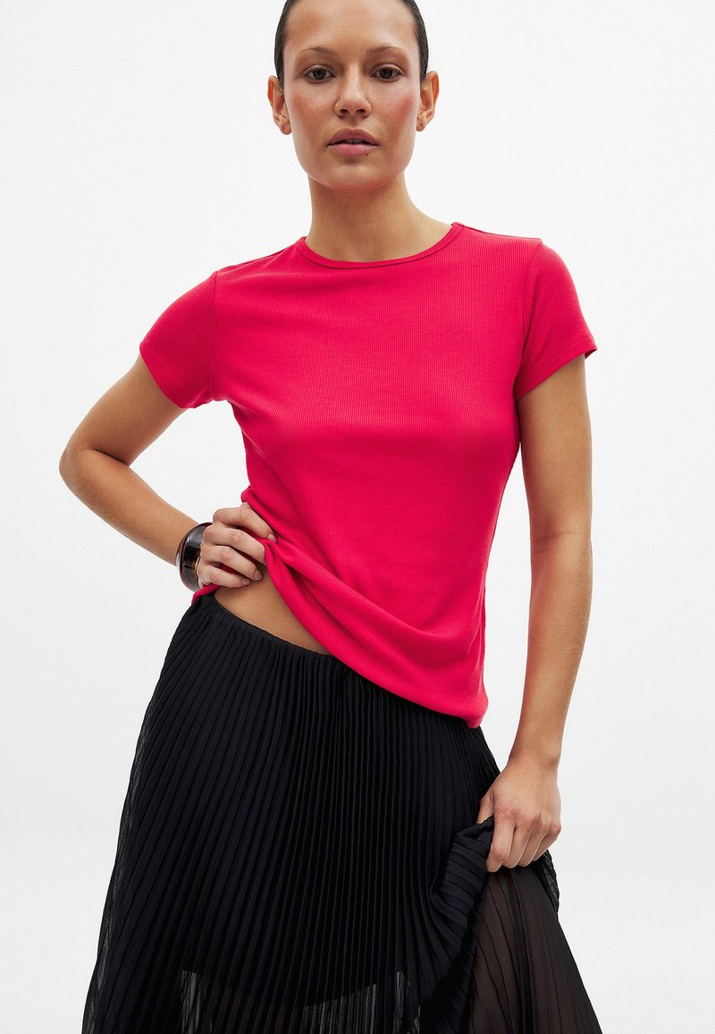 Ryškiai rožinė banguota trumpomis rankovėmis marškinėliai derinami su juoda klostuota sijonu, demonstruojančiu lygią medžiagą ir pritaikytą siluetą.