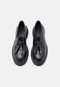 Schwarze Leder-Loafers mit einer klobigen Gummisohle, verziert mit dekorativen Quasten und einem geflochtenen Besatz entlang des Randes. Glatte, glänzende Oberfläche.