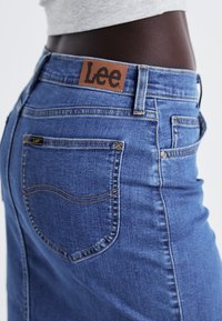 Tumši zili džinsi ar izliekto aizmugures kabatu, brūnā ādas "Lee" uzlīme uz jostas vietas un dzeltenas šuvju akcenti.
