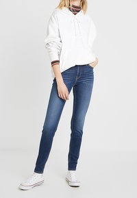 Sudadera blanca con capucha y cordón, combinada con jeans ajustados azules y zapatillas blancas. Diseño sencillo sin patrones visibles.