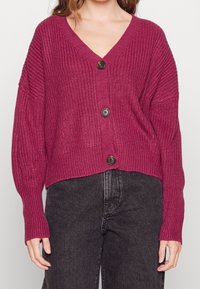 Magenta knitted cardigan with a deep V-neck, long sleeves, and three round dark buttons at the front. Textured material, cropped fit.