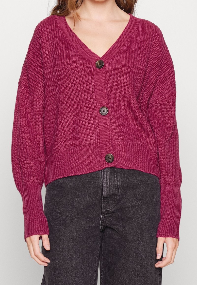 Magenta knitted cardigan with a deep V-neck, long sleeves, and three round dark buttons at the front. Textured material, cropped fit.