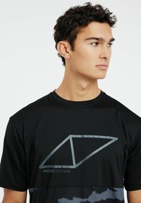 Čierne cyklistické tričko má šedé geometrické logo, krátke rukávy a štruktúrovanú spodnú časť s mriežkovým dizajnom.