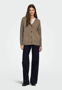 Brauner Strickcardigan mit V-Ausschnitt und sieben schwarzen Knöpfen, kombiniert mit dunkelblauen weiten Hosen, die sichtbare Nahtdetails aufweisen.