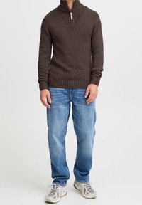Brauner gestrickter Pullover mit hohem Kragen und Reißverschlussdetails, kombiniert mit hellblauen Jeans und grauen Sportschuhen.
