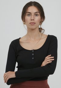 Must mustriline seevallane musternike top, millel on U-kujuline kaelus ja ees kolm hõbedast nuppu, kombineerituna volditud roostepunase seelikuga.