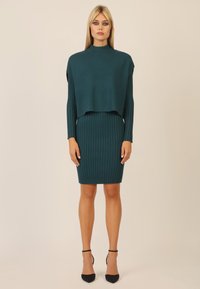 Geripptes türkisfarbenes Kleid mit langen Ärmeln und einem cropped Oberteil, das einen hohen Ausschnitt hat. Schmaler Schnitt, kombiniert mit schwarzen spitzen High Heels.
