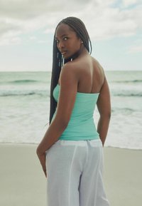 Een vrouw staat bij de zee in een strakke turquoise tanktop en losse witte linnen broeken, die een soepele uitstraling en een relaxte pasvorm tonen.