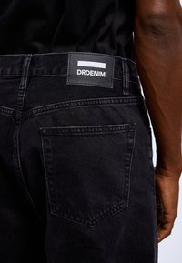 Black denim jeans featuring a rectangular label with "DRDENIM" text. Notable stitching detail and a single back pocket.