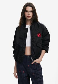 Nadmierne čierne bomber s lesklým povrchom, rebrovaným golierom a červeným vyšívaným detailom. Má dve predné vrecká a zips.