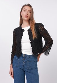 Jachetă scurtă din dantelă neagră, cu decolteu rotund, nasturi de perlă și mâneci lungi transparente, purtată peste un top alb și blugi albaștri.