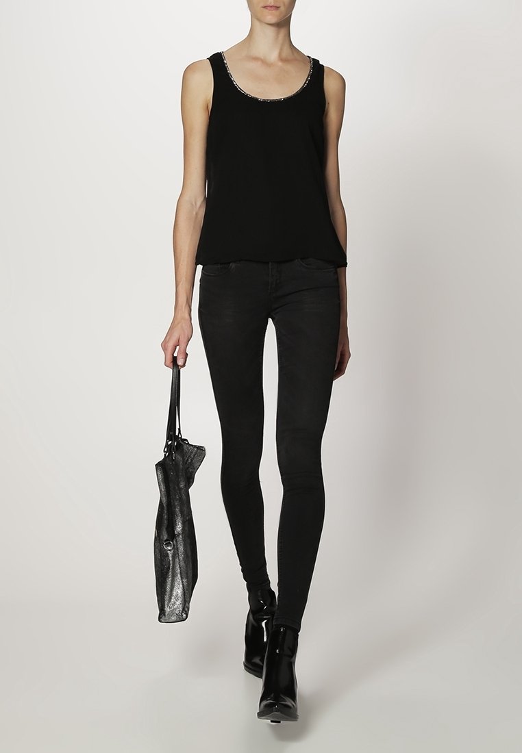 Haut sans manches noir avec un col scintillant, associé à un jean skinny noir et des bottines brillantes, tenant un sac à main métallique foncé.