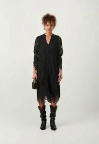 Melna mežģīņu kleita ar garām piedurknēm, ar sarežģītiem izgriezumiem. Apvienota ar melniem zābakiem līdz ceļiem un mazu melnu rokassomu.