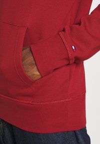 Mano en el bolsillo de un sudadera roja con un pequeño logo de Tommy Hilfiger en el puño de la manga, combinada con jeans de mezclilla azul oscuro.