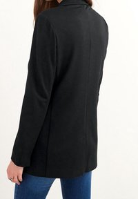 Nainen, jolla on pitkät hihat ja tiukka musta bleiseri, yhdistettynä sinisiin farkkuihin, näkyy selkäpuolelta tasaisella taustalla.