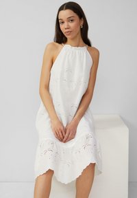 Weißes Neckholder-Kleid mit bestickten Ausschnitten, Wellenkante und leichtem Stoff, getragen von einem Model vor einem neutralen Hintergrund.