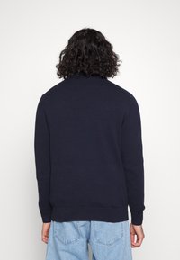 Granatowy sweter dziany z wysokim kołnierzem, ściągaczami przy mankietach i dolnym brzegu. Tył ma gładką fakturę, w zestawieniu z jasnoniebieskimi jeansami.