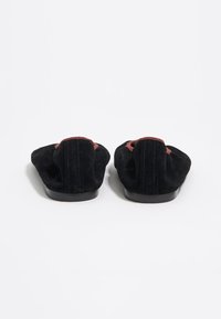 Mocasines de ante negro con una textura suave, que presentan un acento rojo oscuro en el talón y una suela de goma negra de perfil bajo.