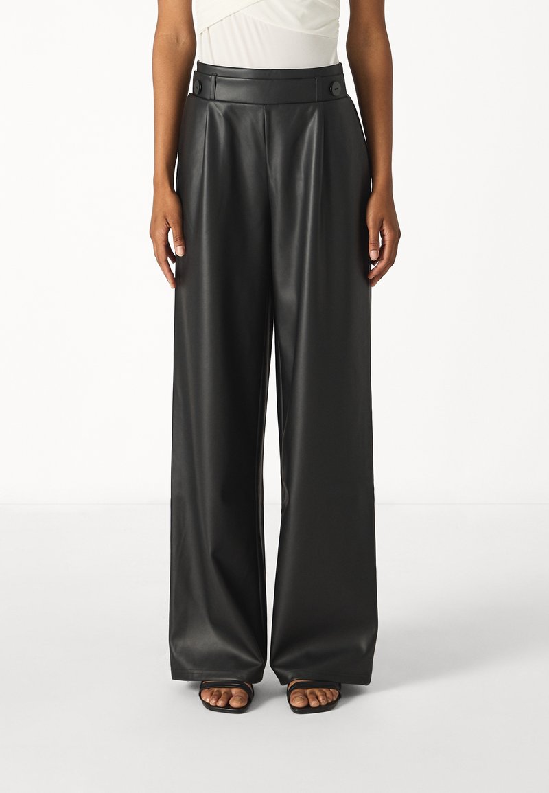 Femme portant un pantalon en cuir noir taille haute à jambes larges avec une ceinture à boutons, associé à des sandales noires à bout ouvert et un haut blanc.