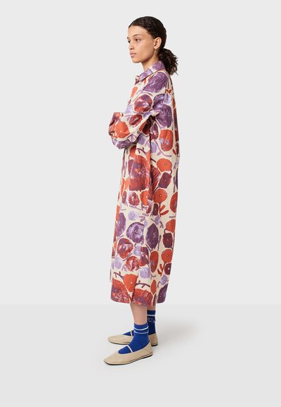 Abito lungo e patterned realizzato in tessuto leggero con grandi motivi floreali rossi e viola, caratterizzato da un colletto e una chiusura frontale con bottoni, indossato con calze blu e scarpe color crema.