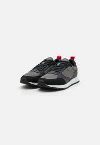 Zwarte en grijze sneakers met een mix van gladde en gestructureerde materialen. Rode accenten op de hiel en tong, witte tussenzool en zwarte buitenzool.