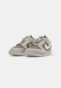 Nike tenisice s glatkom gornjom površinom od sive i smeđe kože, s teksturiranom potplatom i kontrastnim bijelim swoosh logom. Dizajn s vezicama.