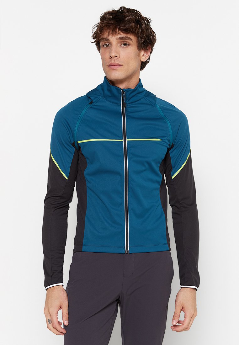Giacca sportiva in teal e nero con zip, presenta strisce riflettenti nella parte anteriore, maniche lunghe e un design aderente. Texture liscia.