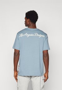 Gai zils kokvilnas T-krekls ar uzrakstu "Los Angeles Dodgers" baltā slīpenā rakstā uz muguras, kā arī etiķete kreisajā pusē netālu no apakšmalas.
