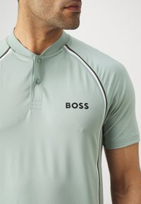 Jasnozielona koszulka polo z krótkim rękawem, z zapinanym dekoltem, czarnym logo "BOSS" oraz kontrastującymi białymi i ciemnozielonymi akcentami wzdłuż szwów.