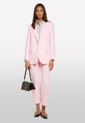 Costume ajusté rose clair avec un revers cranté, poches à rabat et pantalons raccourcis. Associé à une chemise argentée et un sac à main noir.