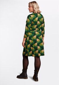 Abito verde con un motivo fogliare giallo e verde scuro. Maniche lunghe e vita aderente, abbinato a stivaletti neri con lacci.