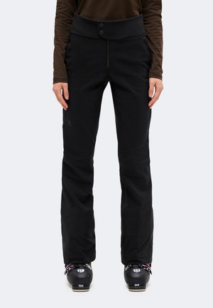 Personne portant un pantalon de ski noir à taille haute avec deux boutons et des chaussures de ski, sur un fond uni.