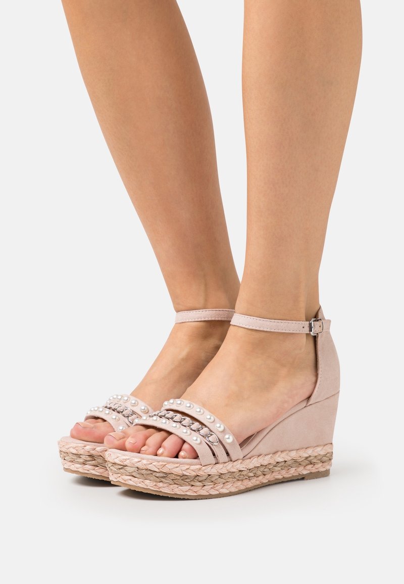 Pieds portant des sandales compensées rose clair avec des brides à la cheville, des ornements en perles et chaînes, et des semelles à plateforme tressées sur fond blanc.