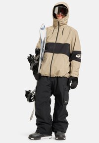 Beżowa i czarna kurtka snowboardowa z kapturem, zapinana na zamek z przodu i z kontrastowym panelem; w połączeniu z czarnymi spodniami snowboardowymi; trzymając snowboard.