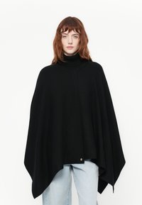 Musta neuleponcho, jossa on korkea korkkiruuvikaulus ja epäsymmetrinen helma. Sisältää koristeellisen metalliosan alaosassa. Yhdistetty vaaleisiin farkkuihin.