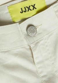Cinturilla de tela blanca con una etiqueta amarilla marcada "JJXX." Botón plateado con diseño grabado y detalle de costura a lo largo de los bordes.