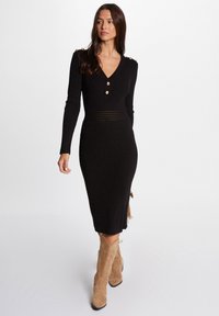 Abito a maglia nero a coste con maniche lunghe, scollo a V, dettagli con bottoni dorati sulle spalle e vita aderente. Abbinato a stivali alti fino al ginocchio di colore tan.