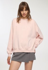 Jasnoróżowa bluza wykonana z miękkiego materiału, z prą�żkowanymi mankietami i dołem, z haftem "SELFLOVE Club" w zaokrąglonym kroju z przodu.