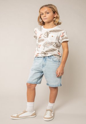 Enfant aux cheveux blonds mi-longs portant un T-shirt blanc à fleurs, un short en denim clair, des chaussettes blanches et des baskets beige-blanc, debout.