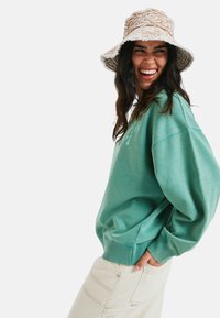 Turkusowy, oversize'owy sweatshirt z prążkowanymi mankietami, zestawiony z jasnobeżowymi spodniami o teksturowej fakturze. Wzorzysty beżowy kapelusz, w stylu bucket, dopełnia cały strój.