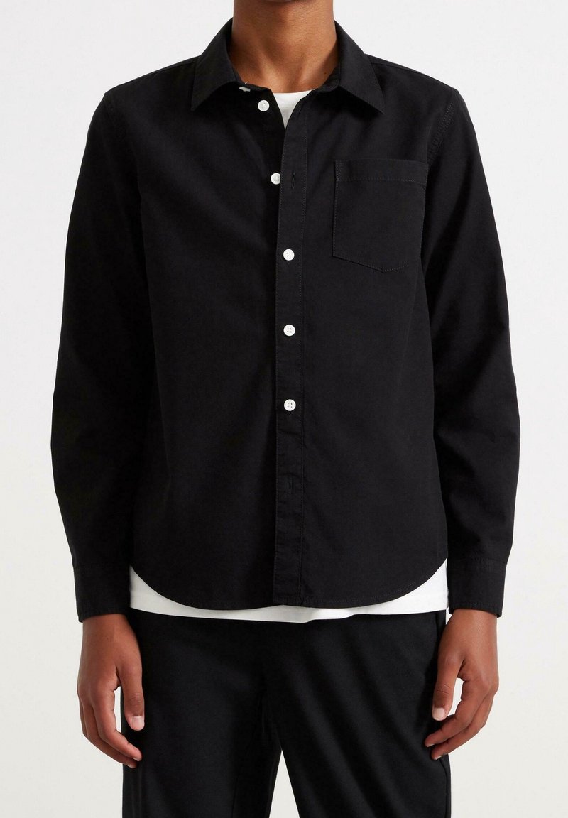 Homme portant une chemise noire à manches longues boutonnée sur un sous-vêtement blanc, associée à un pantalon noir, debout devant un fond blanc.