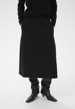 Person wearing a black long skirt, black boots with square toes, and a black long-sleeve top, standing with hands in pockets.