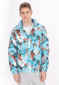 Bloemen windbreaker jas in turquoise, met witte, rode en grijze bloemen. Inclusief een capuchon en een ritssluiting aan de voorkant; gestructureerde stof.