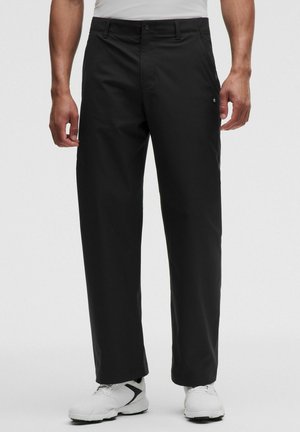 Mann trägt schwarze gerade geschnittene Hose und weiße Sneakers, steht vor schlichtem weißem Hintergrund.