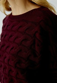 Pull en mailles bourgogne avec un motif en vagues texturé, col rond côtelé et texture de tissu douce. Gros plan sur le design.