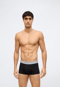 Černé bavlněné boxerky s šedým elastickým páskem se značkou "Calvin Klein." Model má sportovní postavu a stojí na jednolitém pozadí.